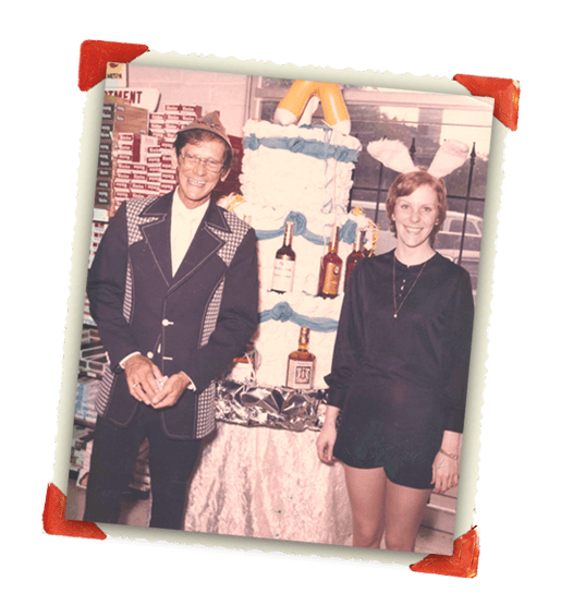 Vintage photograph of two people standing next to a birthday cake decorated with liquor bottles